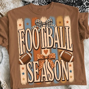 May include: A brown t-shirt featuring a graphic with the text FOOTBALL SEASON in large letters. The design includes two brown footballs, a blue and white checkered bow, an orange polka dot bow, vertical stripes, and small orange and blue stars.