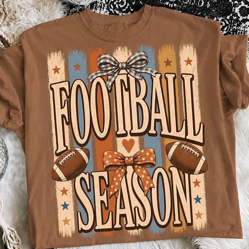 May include: A brown t-shirt featuring a graphic with the text FOOTBALL SEASON in large letters. The design includes two brown footballs, a blue and white checkered bow, an orange polka dot bow, vertical stripes, and small orange and blue stars.