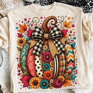 May include: A cream-colored t-shirt featuring a large central graphic of a pumpkin decorated with various patterns including floral, polka dot, chevron, and plaid designs. A black and white buffalo check bow sits atop the pumpkin.