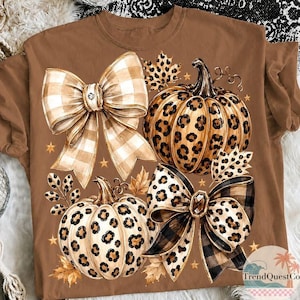 K&ouml;nnte beinhalten: Ein braunes T-Shirt mit einem Grafikdruck von zwei K&uuml;rbissen mit Leopardenmuster, zwei gro&szlig;en dekorativen Schleifen mit Karomuster und Leopardenmuster, herbstlichen Bl&auml;ttern und kleinen goldenen Sternen. Der Text TrendQuest.co ist sichtbar.