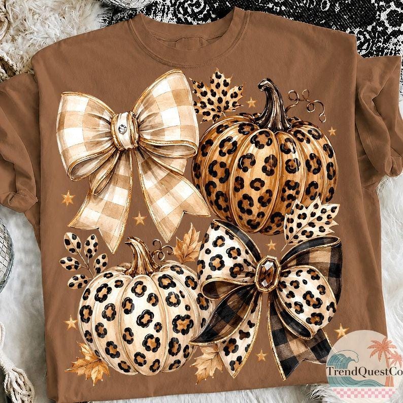 K&ouml;nnte beinhalten: Ein braunes T-Shirt mit einem Grafikdruck von zwei K&uuml;rbissen mit Leopardenmuster, zwei gro&szlig;en dekorativen Schleifen mit Karomuster und Leopardenmuster, herbstlichen Bl&auml;ttern und kleinen goldenen Sternen. Der Text TrendQuest.co ist sichtbar.