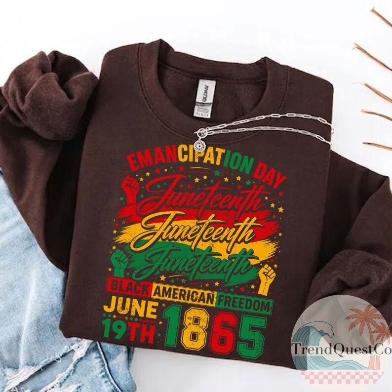 May include: A brown sweatshirt with the text "Emancipation Day Juneteenth" in red, yellow, and green. The design includes the date "June 19th 1865" and the words "Black American Freedom". A silver necklace is draped across the sweatshirt.