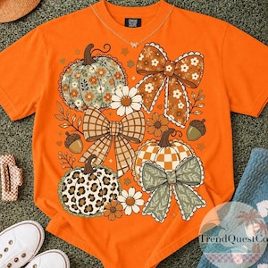 May include: An orange short-sleeved t-shirt featuring a graphic print of three pumpkins, two large bows, and several daisies. The pumpkins display floral, chequered, and leopard patterns. The bows are brown and green with lace-like edges.