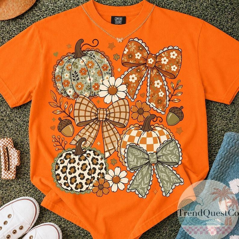 May include: An orange short-sleeved t-shirt featuring a graphic print of three pumpkins, two large bows, and several daisies. The pumpkins display floral, chequered, and leopard patterns. The bows are brown and green with lace-like edges.