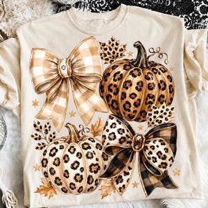 Peut inclure: Un t-shirt couleur crème arborant une impression graphique de deux citrouilles à motifs léopard, deux grands nœuds décoratifs à carreaux et léopard, ainsi que plusieurs feuilles d'automne et petites étoiles dorées éparpillées sur le devant.