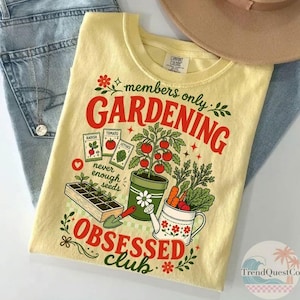 Puede incluir: Una camiseta amarilla clara con un tema de jardiner&iacute;a. La camiseta presenta las palabras "members only GARDENING OBSESSED club" en rojo y verde, junto con ilustraciones de verduras, sobres de semillas y una regadera. La camiseta se combina con unos vaqueros azules y un sombrero marr&oacute;n.