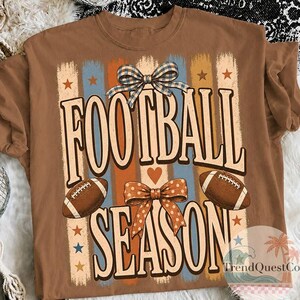 May include: A brown t-shirt featuring a graphic print with the text FOOTBALL SEASON in large letters. The design includes two brown footballs, a blue and white checkered bow, an orange polka dot bow, and vertical stripes in blue and orange.