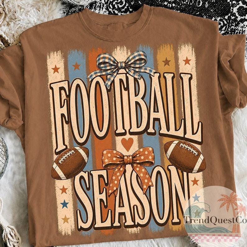 May include: A brown t-shirt featuring a graphic print with the text FOOTBALL SEASON in large letters. The design includes two brown footballs, a blue and white checkered bow, an orange polka dot bow, and vertical stripes in blue and orange.