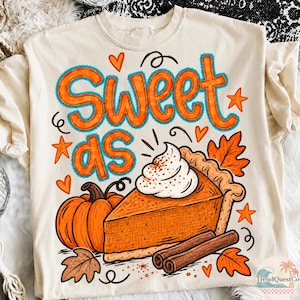 May include: A cream-colored t-shirt featuring a graphic print with the text "SWEET AS" in orange letters with blue outlines. The design includes a pumpkin, a slice of pumpkin pie topped with whipped cream, cinnamon sticks, and autumn leaves.