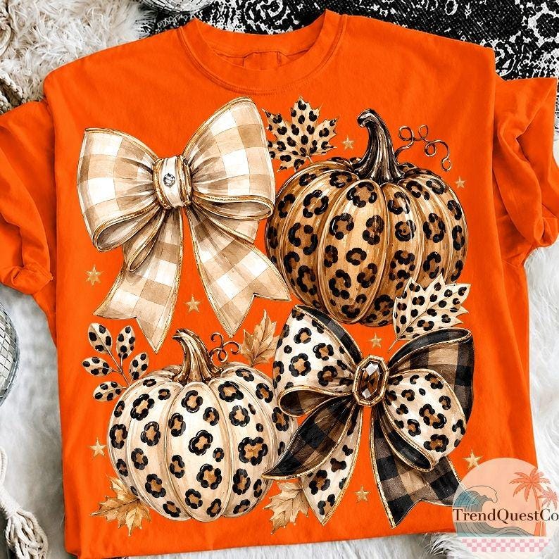 K&ouml;nnte beinhalten: Ein leuchtend orangefarbenes T-Shirt mit einem Grafikdruck von zwei K&uuml;rbissen und zwei gro&szlig;en Schleifen. Die K&uuml;rbisse haben ein beigefarbenes und braunes Leopardenmuster. Eine Schleife ist beige-wei&szlig; kariert, die andere beige-schwarz kariert mit Leopardenakzenten.