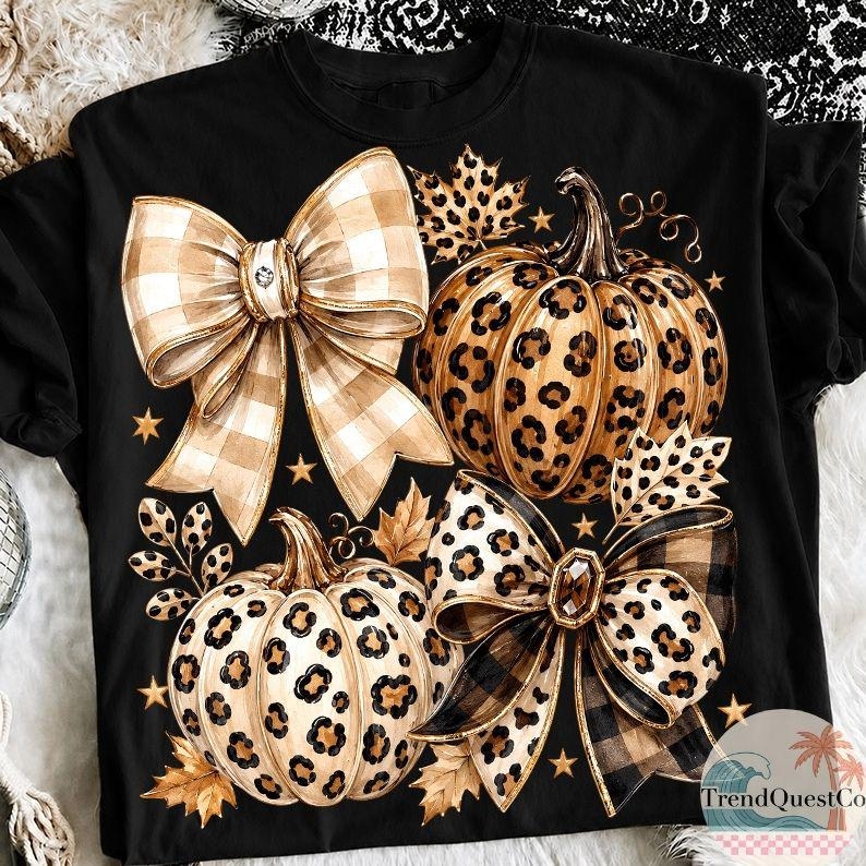 K&ouml;nnte beinhalten: Ein schwarzes T-Shirt mit einem Grafikdruck von zwei K&uuml;rbissen und zwei gro&szlig;en Schleifen. Die K&uuml;rbisse haben ein Leopardenmuster, w&auml;hrend die Schleifen ein beigefarbenes und braunes Karomuster aufweisen. Kleine goldene Sterne und Herbstbl&auml;tter sind &uuml;ber das Motiv verteilt.