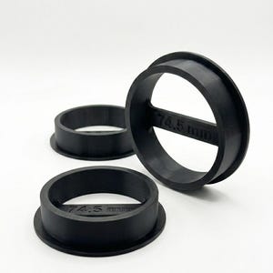 Op de afbeelding: Drie zwarte cirkelvormige ringen met een platte basis en een horizontale dwarsbalk in het midden. Elke ring is voorzien van de tekst 74.5 mm in reliëf op de centrale balk. De objecten zijn gegroepeerd en hebben een matte afwerking.