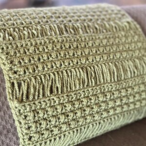 May include: A close-up of a green crocheted pillow with a textured pattern. The pillow is placed on a brown textured surface.