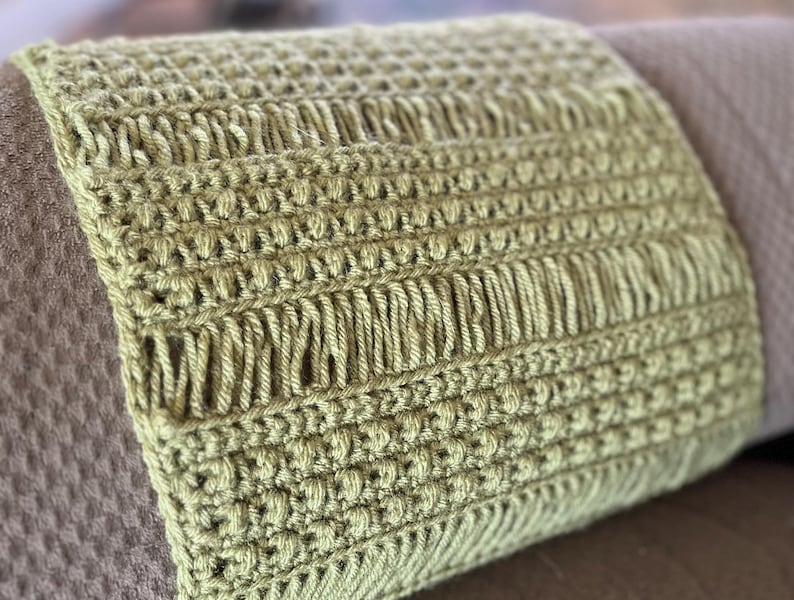 May include: A close-up of a green crocheted pillow with a textured pattern. The pillow is placed on a brown textured surface.