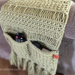 May include: A green crocheted couch armrest with two pockets for remotes or other small items. The armrest has a fringe on the bottom and a brown leather tag with the text "Yarnnovations.com".