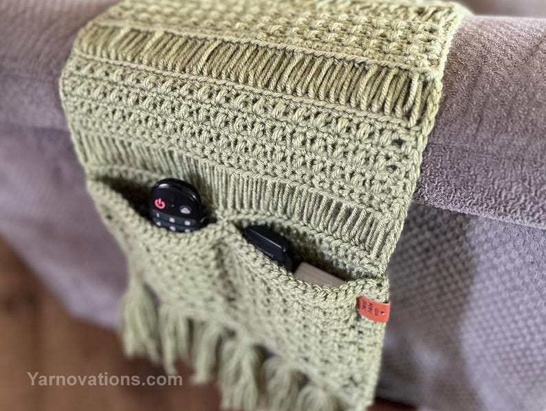 May include: A green crocheted couch armrest with two pockets for remotes or other small items. The armrest has a fringe on the bottom and a brown leather tag with the text "Yarnnovations.com".