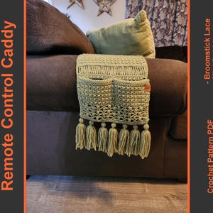 May include: A crocheted green remote control caddy with tassels hanging from the bottom. The caddy has two pockets and is attached to the arm of a couch.