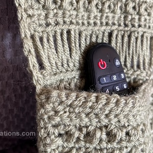 May include: A close-up of a crocheted pocket with a black remote control inside. The pocket is made with a beige yarn and has a textured pattern. The remote control has a power button and number buttons.