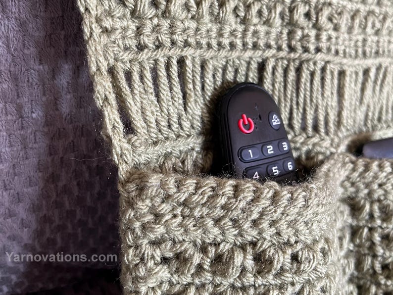 May include: A close-up of a crocheted pocket with a black remote control inside. The pocket is made with a beige yarn and has a textured pattern. The remote control has a power button and number buttons.