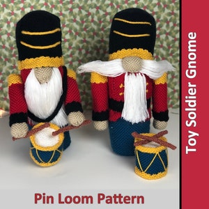 May include: Two crocheted toy soldier gnomes with red, blue, and yellow accents. Each gnome is holding a small drum. The gnomes are standing on a white surface. The image includes the text "Pin Loom Pattern" and "Toy Soldier Gnome".