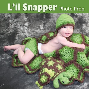 May include: A crochet turtle photo prop set for babies. The set includes a hat, cape, diaper cover, blanket, rattle, and toy. The set is green and brown and features a crocheted turtle design.