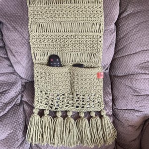 May include: A crocheted green remote control holder with two pockets and tassels. The holder is attached to the arm of a chair.