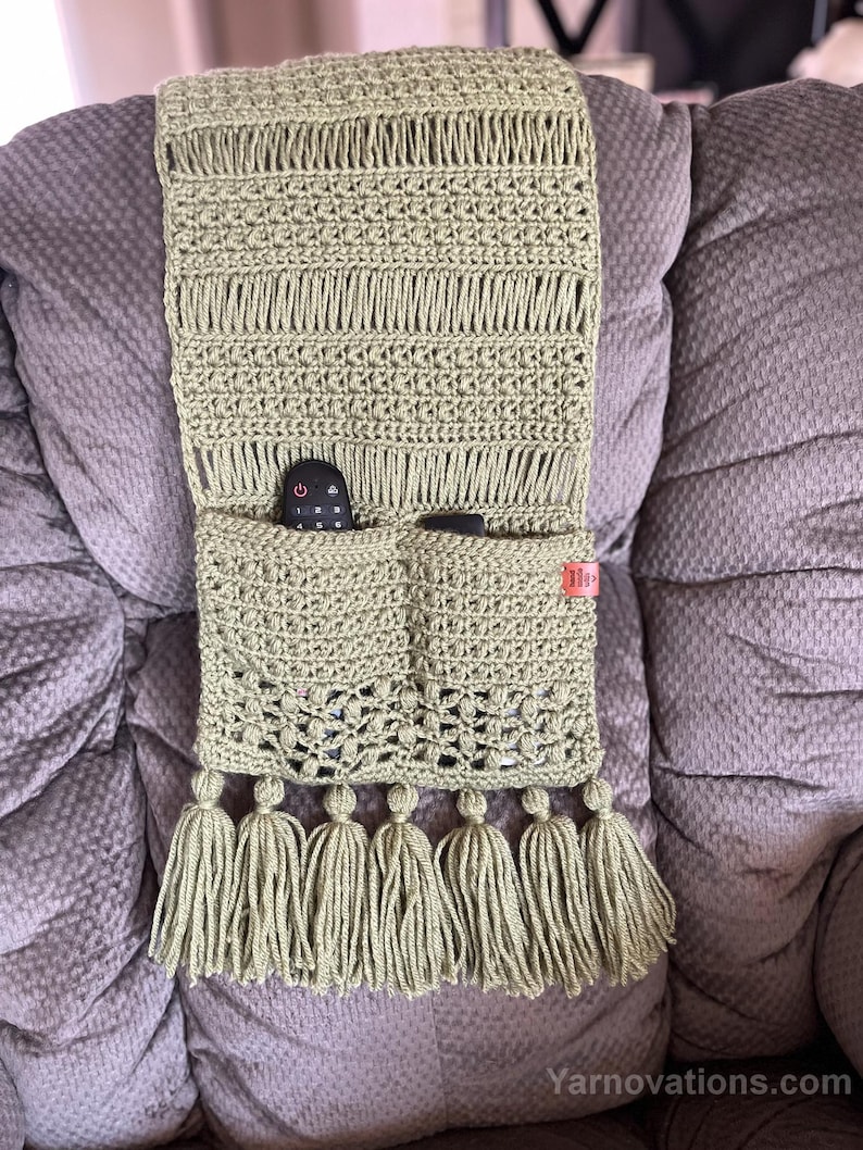 May include: A crocheted green remote control holder with two pockets and tassels. The holder is attached to the arm of a chair.