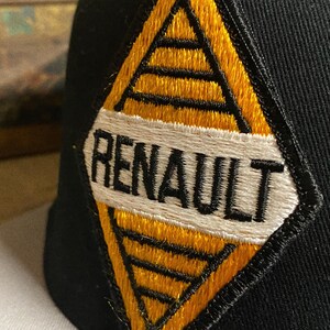 Op de afbeelding: Zwarte baseballcap met een geel en zwart geborduurd embleem. Het embleem toont het woord "RENAULT" in zwarte letters op een witte achtergrond, omgeven door een geel en zwart geometrisch ontwerp.