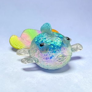 May include: A translucent, round fish figurine featuring a glittery blue and iridescent body with black eyes. The fish has prominent side fins and a large, flowing tail fin that transitions from yellow to pink and green hues in a gradient pattern.