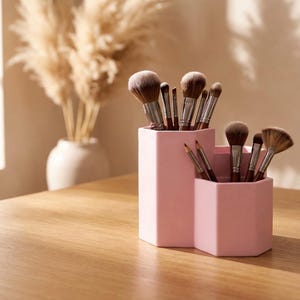 Puede incluir: Un soporte rosa geométrico para brochas de maquillaje lleno de varios pinceles. El soporte está sobre una mesa de madera, con un jarrón de flores secas en el fondo. La escena está iluminada por luz natural.