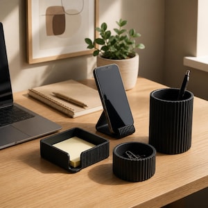 Op de afbeelding: Een moderne bureau-opstelling met een laptop, smartphone op een standaard en bijpassende zwarte bureau-accessoires. Inclusief een pennenhouder, een klein bakje en een notitieblok met een pen. De items zijn geplaatst op een licht houten bureau.