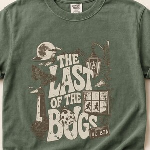 Puede incluir: Una camiseta de manga corta color verde salvia con un estampado gr&aacute;fico marr&oacute;n que incluye el texto THE LAST OF THE BUGS. El dise&ntilde;o muestra un faro, una polilla, una mariquita, un farol, una ventana con dos figuras y una matr&iacute;cula 4C B3A.