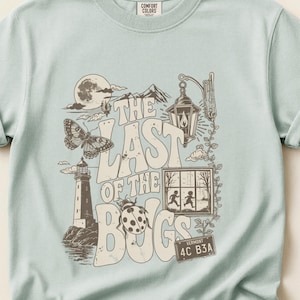 Puede incluir: Una camiseta de manga corta en color verde salvia claro con un estampado gr&aacute;fico en marr&oacute;n oscuro que incluye el texto THE LAST OF THE BUGS. El dise&ntilde;o muestra un faro, una mariposa, una mariquita, un farol, una ventana con dos figuras y una matr&iacute;cula de Vermont.