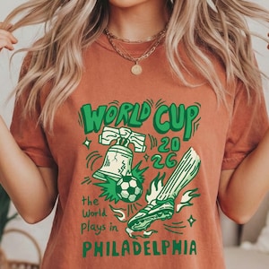 May include: A terracotta-colored short-sleeve t-shirt featuring a green graphic print that includes a Liberty Bell, a soccer ball, and a soccer cleat. The text reads World Cup 2026 the World plays in PHILADELPHIA in a stylized green font.