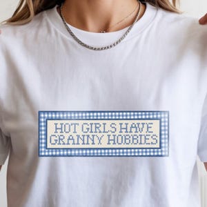 May include: White t-shirt with the text "HOT GIRLS HAVE GRANNY HOBBIES" in a blue and white gingham pattern. The text is in a cross-stitch style, and the shirt is a classic crew neck.