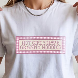 May include: White t-shirt with the text "HOT GIRLS HAVE GRANNY HOBBIES" in a pink and white gingham-style frame. The text is in a cross-stitch style font. The shirt is a casual, everyday wear item.