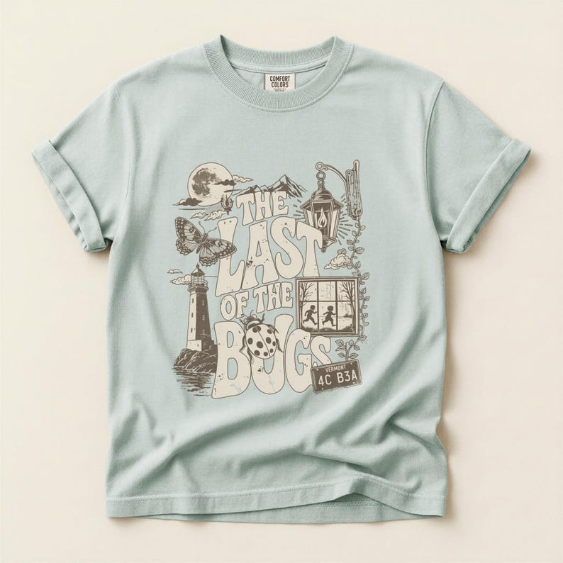 Puede incluir: Una camiseta de manga corta en color verde salvia claro con un estampado gr&aacute;fico en marr&oacute;n oscuro que incluye el texto THE LAST OF THE BUGS. El dise&ntilde;o muestra un faro, una mariposa, una mariquita, un farol, una ventana con dos figuras y una matr&iacute;cula de Vermont.