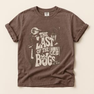 Puede incluir: Una camiseta marr&oacute;n de manga corta con un estampado gr&aacute;fico que incluye el texto THE LAST OF THE BUGS. El dise&ntilde;o muestra una luna, un faro, una linterna, una mariquita, una ventana con dos figuras y una matr&iacute;cula con VERMONT 4C B3A.