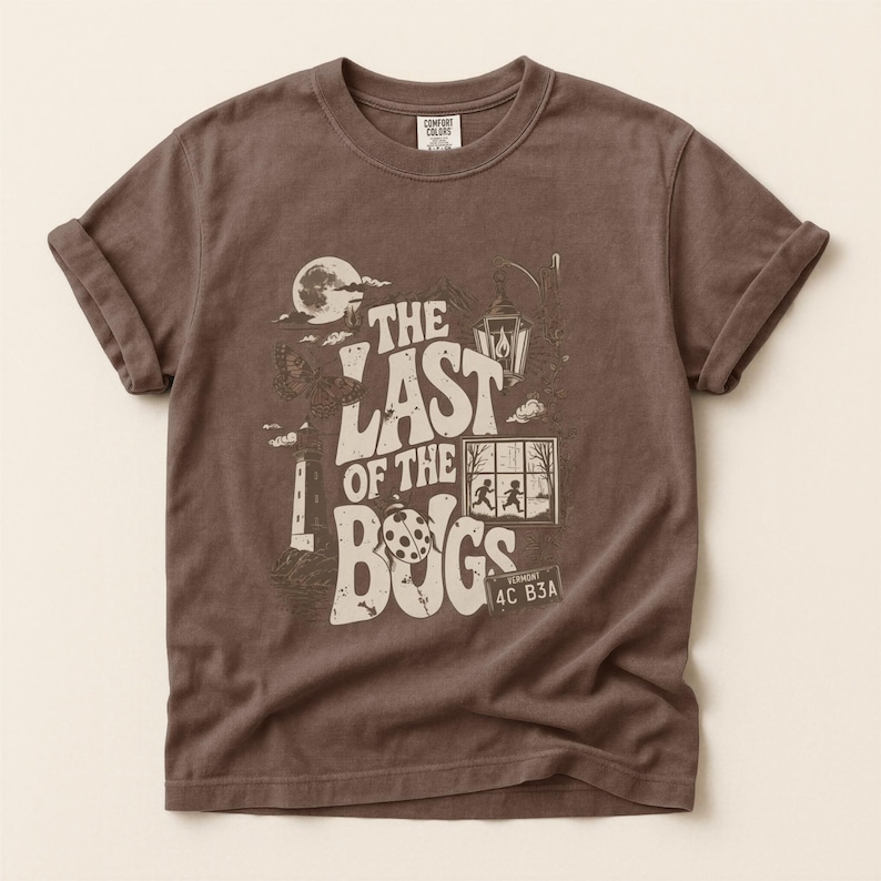 Puede incluir: Una camiseta marr&oacute;n de manga corta con un estampado gr&aacute;fico que incluye el texto THE LAST OF THE BUGS. El dise&ntilde;o muestra una luna, un faro, una linterna, una mariquita, una ventana con dos figuras y una matr&iacute;cula con VERMONT 4C B3A.