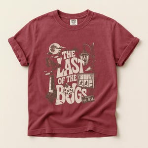 Puede incluir: Una camiseta de manga corta de color rojo oscuro con un estampado gr&aacute;fico en crema y marr&oacute;n. El dise&ntilde;o incluye una luna, un faro, un farol, una mariquita y una ventana con dos figuras, junto con el texto THE LAST OF THE BUGS y una matr&iacute;cula de Vermont.