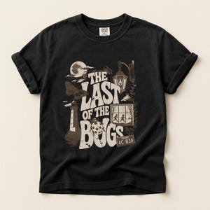 Puede incluir: Una camiseta negra de manga corta con un estampado gr&aacute;fico que incluye el texto THE LAST OF THE BUGS. El dise&ntilde;o muestra un faro, una polilla, una luna, un farol, una ventana con dos siluetas y una matr&iacute;cula de Vermont con el texto 4C B3A.