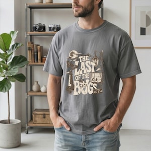 Puede incluir: Una camiseta gris carb&oacute;n de manga corta con un estampado gr&aacute;fico que incluye el texto THE LAST OF THE BUGS. El dise&ntilde;o muestra un faro, una luna, una linterna, una mariquita y una ventana con dos siluetas, en tonos crema y marr&oacute;n.