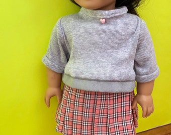 May include: A doll wearing a gray sweater with a pink heart detail, a plaid skirt, and silver shoes with pink flowers.