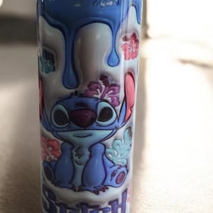 May include: A white tumbler featuring a blue cartoon character with large eyes, pink ears, and a floral crown. The design includes blue drips and the word "Stitch" in blue and pink.