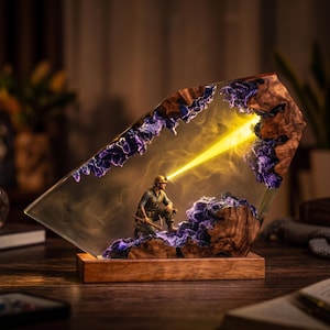 May include: A decorative lamp featuring a miniature miner figurine kneeling inside a clear resin block with natural wood accents and purple crystalline structures. A bright yellow light beam emanates from the miner's headlamp, illuminating the interior.