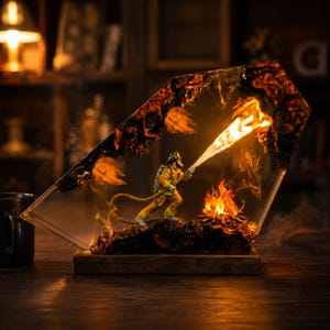 Könnte beinhalten: Eine dekorative Harzlampe mit einem Miniatur-Feuerwehrmann in gelber Schutzausrüstung, der einen Schlauch hält, der helles Licht ausstrahlt. Die Szene ist in einen klaren, kantigen Harzblock mit dunklen, verkohlten Kanten und orangefarbenen Flammen eingebettet.