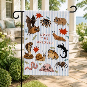 May include: A garden flag with a blue and white striped background featuring cartoon illustrations of various animals, including a bat, spider, mouse, snake, snail, frog, and worm. The text "LOVE THE UNLOVED" is in the center.