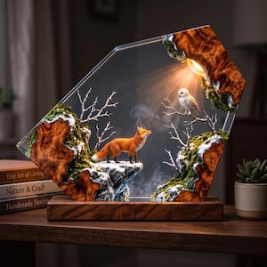 Peut inclure: Une lampe décorative avec un cadre en résine transparente en forme de losange renfermant une scène de forêt miniature avec un renard, un hibou, des branches nues, de la mousse verte et des touches de neige blanche sur un socle en bois.