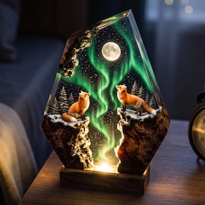 May include: A decorative lamp featuring a resin-encased scene of two orange foxes standing on snowy, dark wood cliffs. The background displays a vibrant green aurora borealis, a bright full moon, and a starry night sky above pine trees.