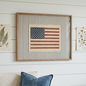Puede incluir: Una pieza de arte mural rectangular enmarcada que presenta una impresi&oacute;n centrada de la bandera de los Estados Unidos con una paleta de colores rojo, blanco y azul apagados. La bandera se sit&uacute;a sobre un fondo de rayas verticales negras en papel crema.